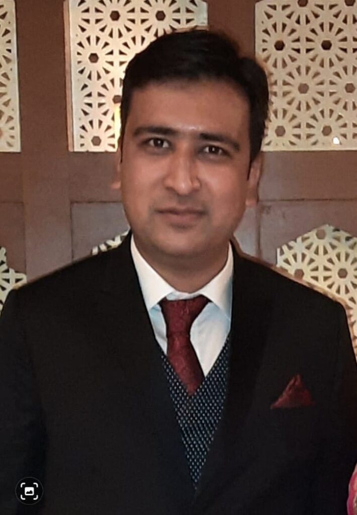 Well-dressed Indian man in a black suit, white shirt, and maroon tie with pocket square, posing confidently indoors against a decorative wooden panel background.