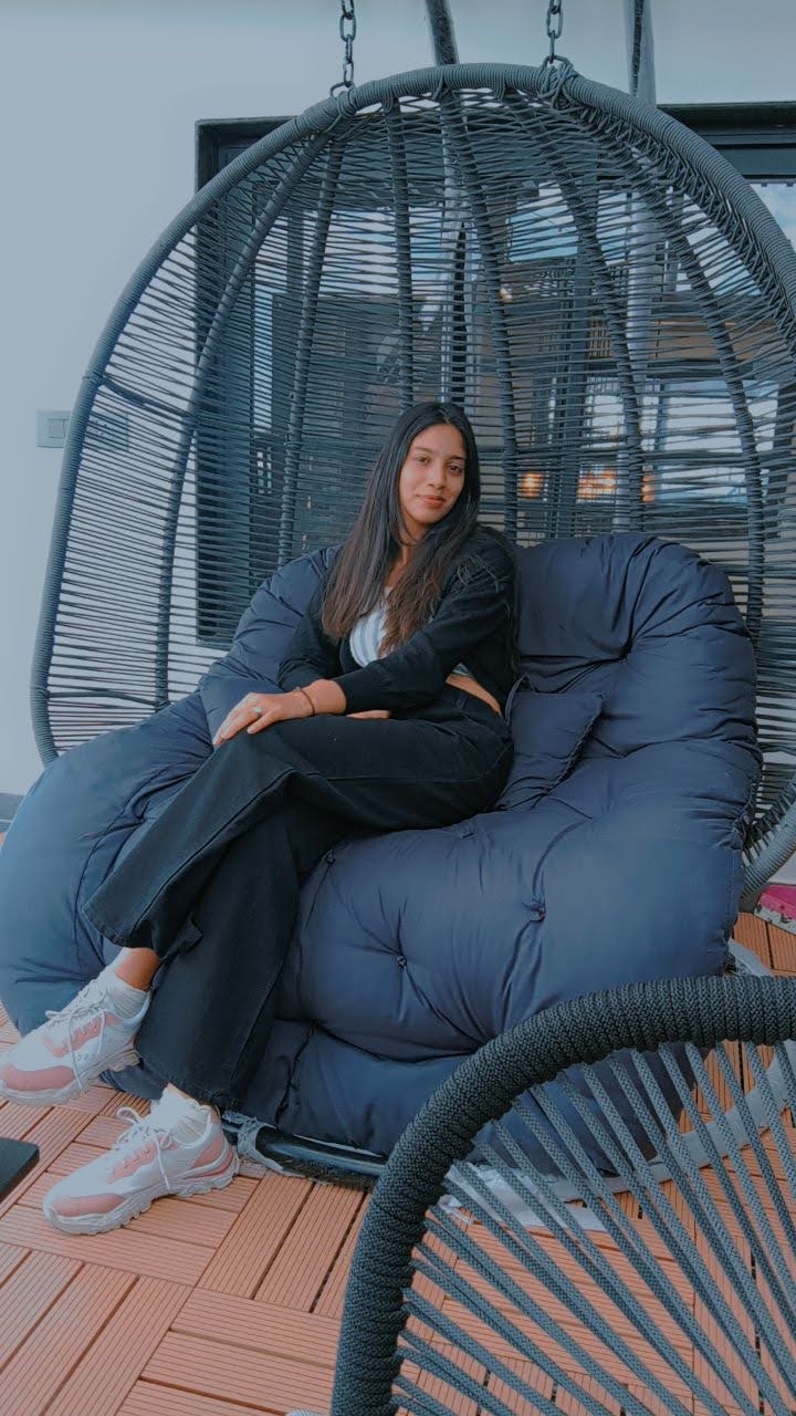 A young woman sitting comfortably on a large cushioned swing chair, dressed in casual black attire with sneakers, posing calmly indoors