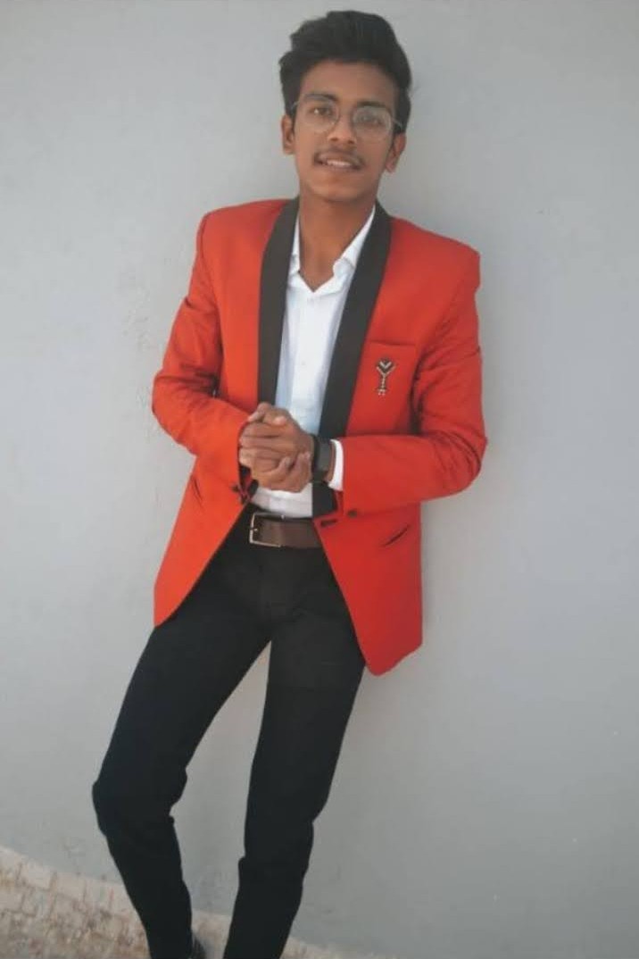 Young Indian man wearing a stylish red blazer with black lapel, white shirt, and black trousers, posing confidently against a plain wall.