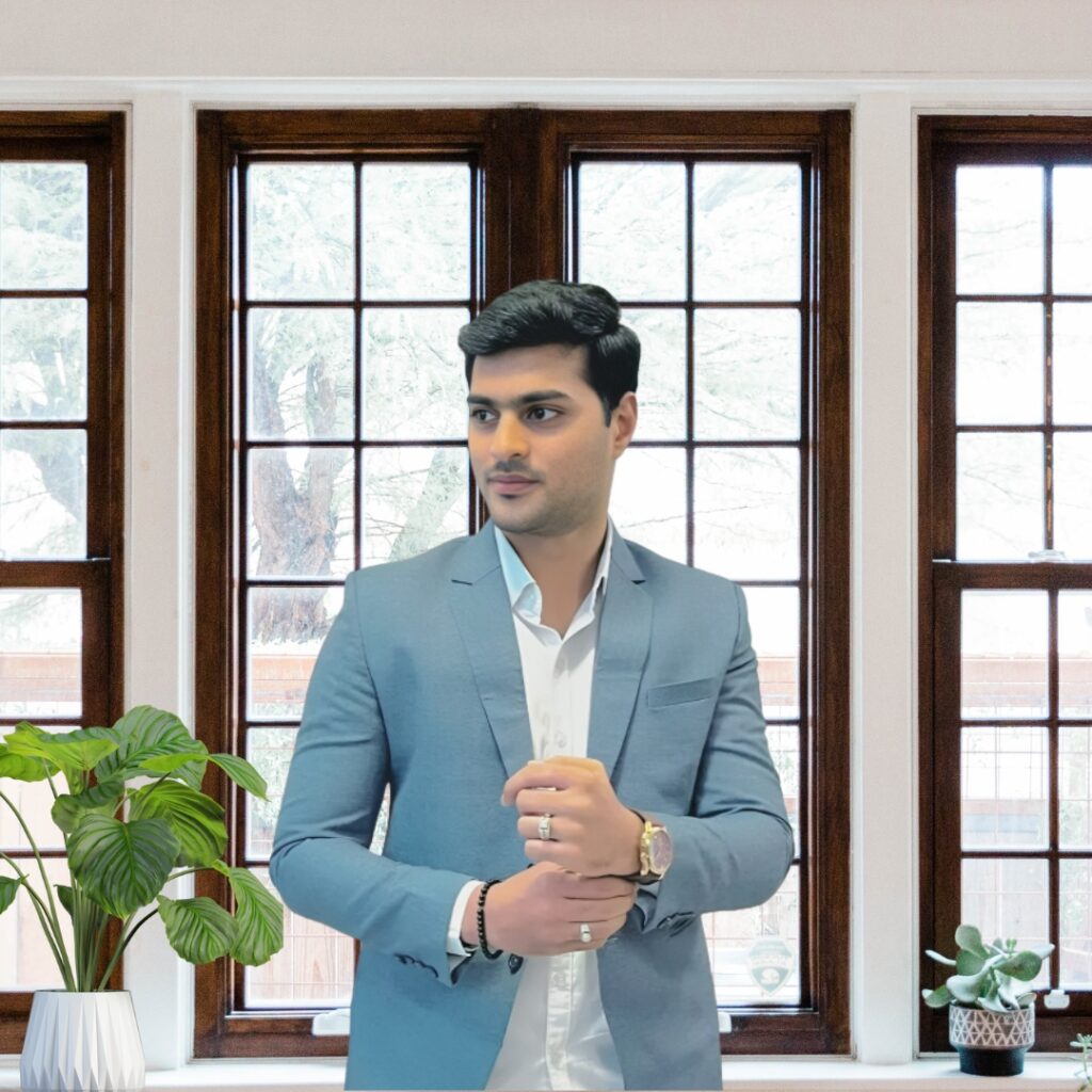 Minimalist professional portrait of a man in a light blue suit adjusting his cuff in front of large windows with indoor plants.