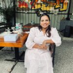 Woman in a white embroidered dress smiling while seated at a café with graffiti art in the background.