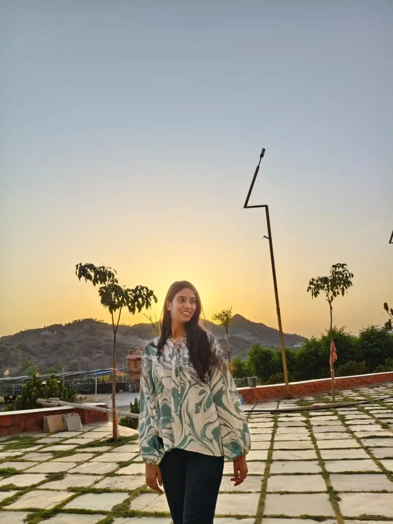 Woman standing outdoors during sunset with hills in the background, smiling and enjoying a calm golden-hour moment in nature.
